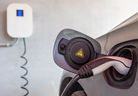 A close-up of an electric car being charged, showing the charging plug inserted into the car’s port with a high-voltage warning symbol. A wall-mounted charging station is visible in the background.