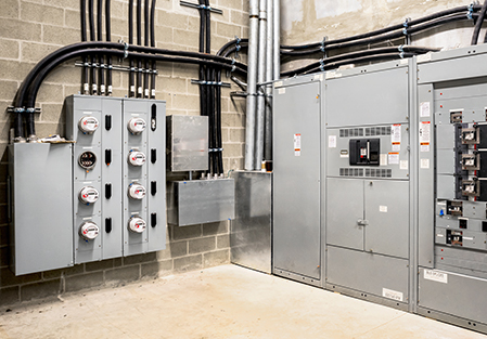 A utility room with multiple electric meters mounted on a wall, several grey electrical panels, conduit pipes, and black cables, all set against a cinder block wall with a concrete floor.