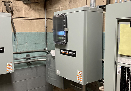 A gray Generac Industrial Power electrical panel is mounted on a wall in a utility room, surrounded by pipes, wires, and other electrical equipment. A transparent cover reveals switches inside the panel.