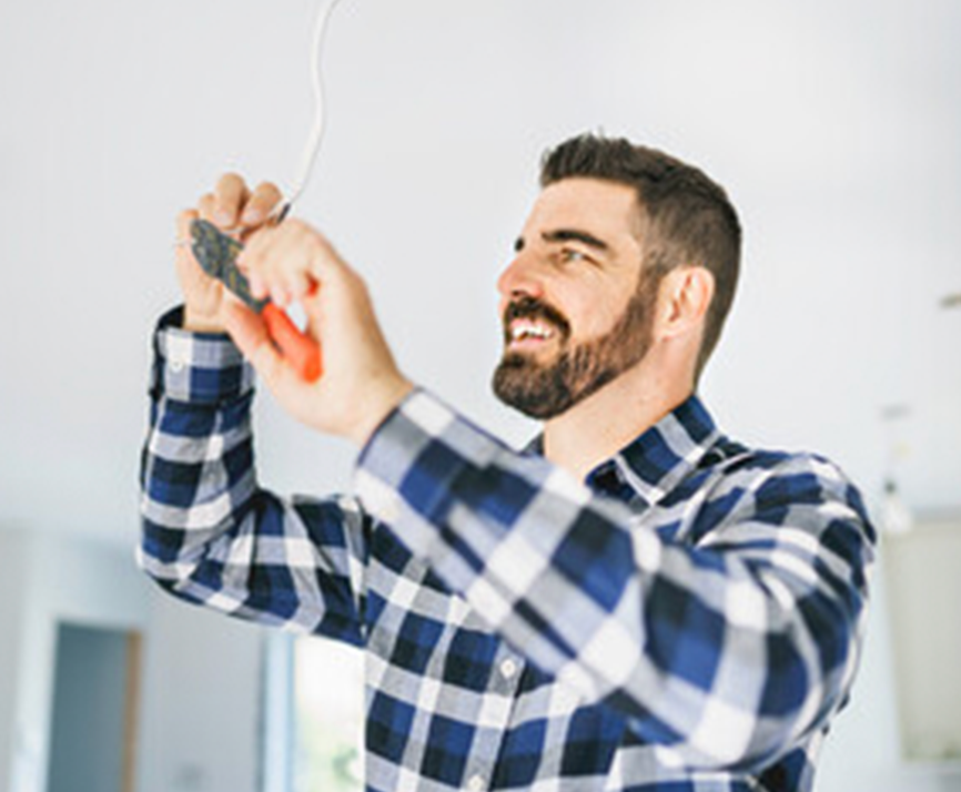 A man in a plaid shirt smiles while using pliers to work on an electrical wire on the ceiling.