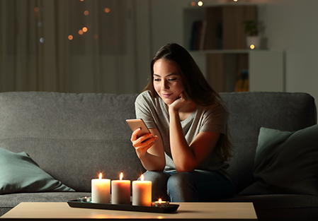 A woman sits on a couch in a dimly lit room, smiling as she looks at her smartphone. Several lit candles on a tray illuminate the scene, creating a cozy atmosphere.