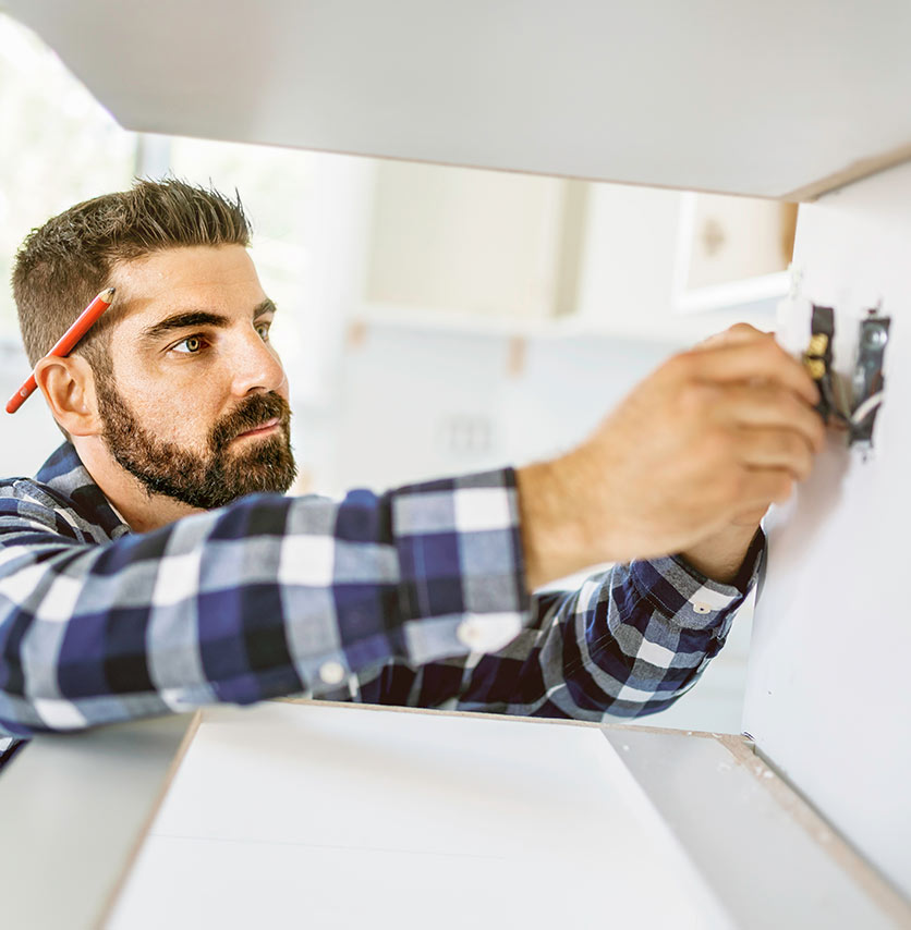 A man with a pencil behind his ear, wearing a plaid shirt, installs or repairs an electrical outlet on a wall, focusing intently on his work.