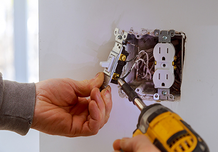 A person uses a power drill to install or remove a light switch next to an electrical outlet in a wall, exposing wires and electrical components.