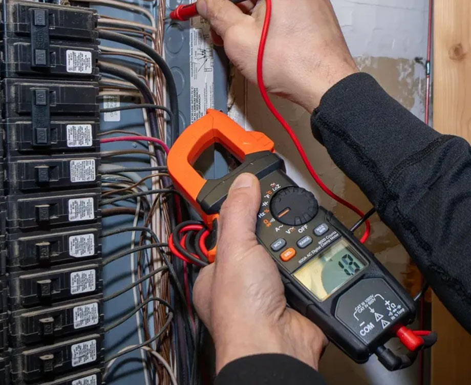 A person uses a digital clamp multimeter to test electrical connections inside a breaker panel, holding the probes against wires and measuring voltage. The panel contains multiple circuit breakers and organized wiring.