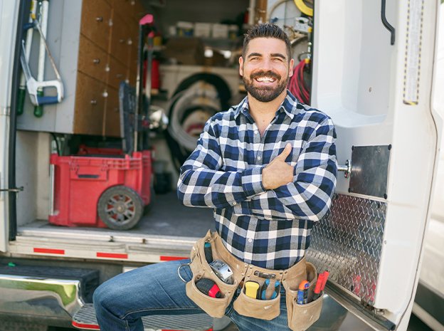 A smiling man in a plaid shirt with a tool belt stands by an open work van filled with tools and equipment, arms crossed and looking confident, suggesting he is a handyman or technician.